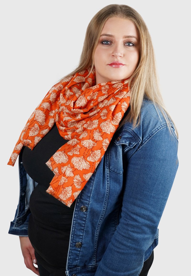 Oranje sjaal met een wit bloemenpatroon, om de nek gedrapeerd, gecombineerd met een spijkerjack. Zacht, lichtgewicht materiaal met een gestructureerde afwerking.