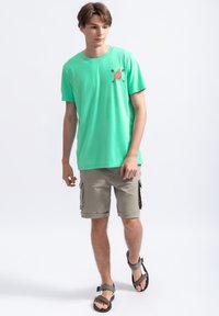 Groene katoenen t-shirt met krabafbeelding, gedragen met beige cargoshorts met zijzakken en bruine sandalen met verstelbare banden.