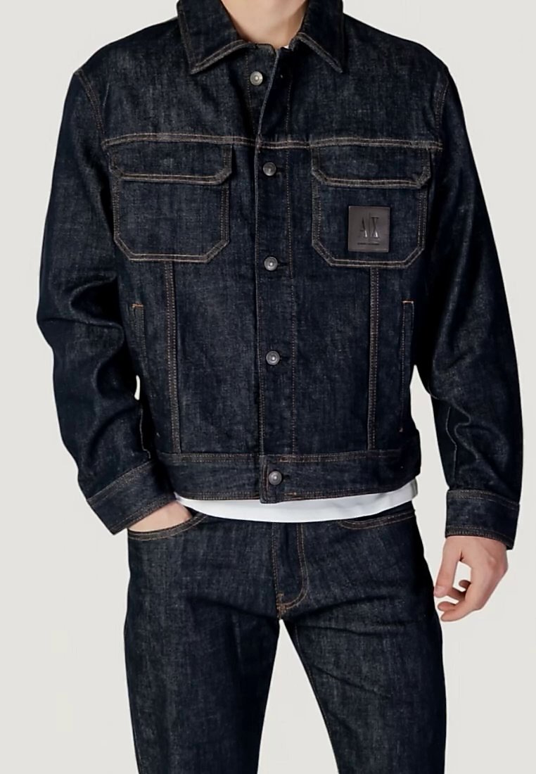 Homme portant une veste en jean bleu marine et un jean, avec la veste boutonnée et les mains en partie dans les poches.