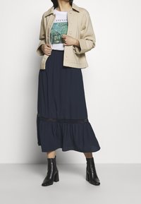 Beige bomullsväst med knappar, vit grafisk t-shirt, marinblå lager-på-lager kjol med spetskant, och svarta ankelskor.