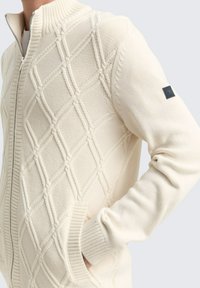 Cremefarbener Reißverschluss-Pullover mit strukturiertem Rautenmuster, geripptem Kragen und Bündchen sowie einem kleinen Akzentlogo am Ärmel.
