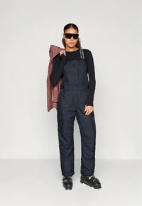 Macacão de esqui azul-marinho com isolamento térmico, alças ajustáveis, fecho de correr frontal e vários bolsos. Modelo a segurar um casaco rosa, a usar óculos de sol e botas de esqui.