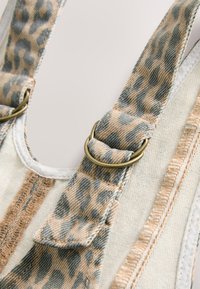 Bretelles réglables en tissu beige et gris à motif léopard avec anneaux métalliques sur un vêtement en denim clair.