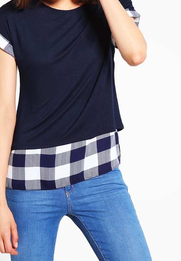 Kvinna i en marinblå lagerblus med en svart-vit rutig skjorta under och blåjeans, arm böjd vid huvudet, vit bakgrund.