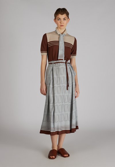 Braun-beige Oberteil mit kurzen Ärmeln und Kragen; hellblaue Rock mit geometrischem Muster und Kordelzugbund; braune Rutschsandalen.