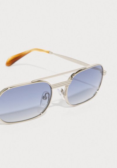 Metāla saulesbrilles ar sudraba apdari, zilām gradienta lēcām un brūniem akcentiem uz tempļiem. Taisnstūra forma un regulējami deguna spilventiņi.