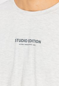 Gaiši pelēks T-krekls, kas izgatavots no mīkstas auduma, ar apaļu kakla izgriezumu un melniem uzrakstiem: "STUDIO EDITION ALPHA INDUSTRIES INC."