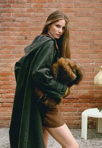 Giovane donna in un cappotto oversize verde scuro e gonna marrone tiene una grande borsa pelosa contro uno sfondo di muro di mattoni.