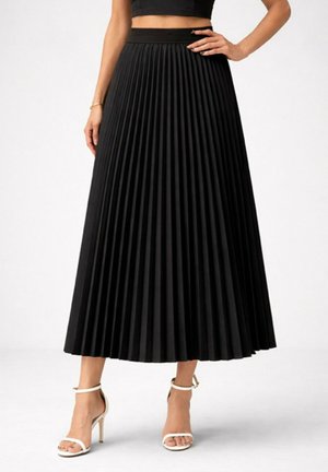 Jupe midi plissée noire taille haute portée avec un crop top noir et des sandales blanches à talons hauts avec bride à la cheville sur fond blanc.