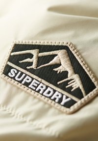 Toppa in tessuto beige con una forma di diamante nera e un logo in rilievo che mostra "SUPERDRY" in bianco e grafiche di montagne testurizzate.