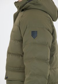 Oliven grøn polstret jakke med hætte, med en tekstureret overflade, et logo patch på venstre ærme, og trykknapdetaljer.