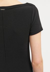Chemise noire à manches courtes en tissu texturé. Encolure arrondie et couture centrale dans le dos. Étiquette de marque en métal près de l'encolure.