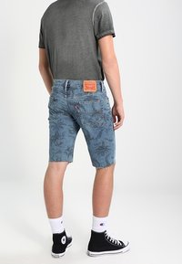 Denim shorts in light blue with a leaf pattern, frayed hem, and leather patch on the waistband. Paired with a gray t-shirt and black sneakers.