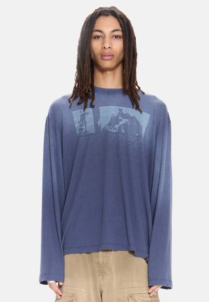 Persoon met lange dreadlocks draagt een loszittend blauw shirt met lange mouwen met vervaagde grafische prints en beige cargobroek tegen een witte achtergrond.