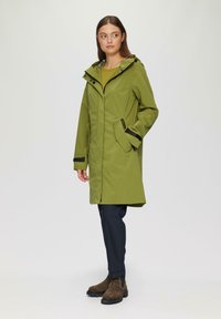 Zielony wodoodporny płaszcz z kapturem, z przednim zamkiem błyskawicznym, kieszeniami po bokach i czarnymi akcentami. W zestawie z ciemnymi spodniami i botkami za kostkę.