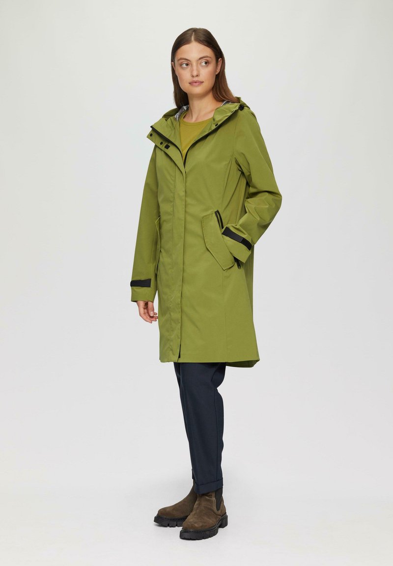 Zielony wodoodporny płaszcz z kapturem, z przednim zamkiem błyskawicznym, kieszeniami po bokach i czarnymi akcentami. W zestawie z ciemnymi spodniami i botkami za kostkę.