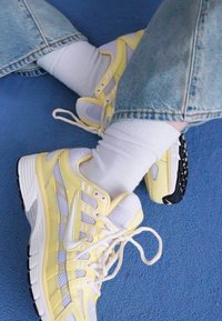 Gele en witte sneakers met mesh-panelen, een textuurzool en veters, gecombineerd met witte sokken en lichtblauwe jeans.