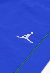 Zils audums ar gludu tekstūru, kurai ir balts izšūts Jumpman logo. Zaļa akcenta līnija šķērsām iet gar malu.