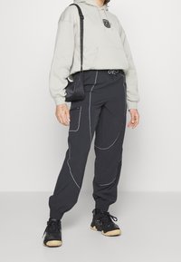 Zwarte broek met witte stiksels, elastische manchetten en zijzakken, gecombineerd met een lichtgrijze hoodie met een logo aan de voorkant en een trekkoord.