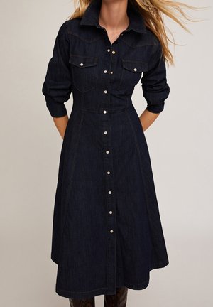 Femme portant une robe midi en jean foncé boutonnée, manches longues, poches poitrine et col montant, debout devant un fond uni.