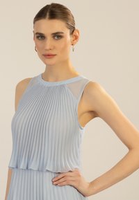 Robe plissée bleu clair avec des accents transparents sur les épaules. Tissu doux, sans manches, présentant des plis verticaux texturés et un ourlet superposé.