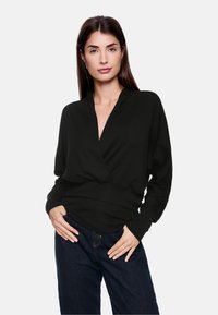 Schwarzes Wickeloberteil mit langen Ärmeln, tiefer V-Ausschnitt und taillierter Passform. Weicher Stoff mit leichtem Glanz, kombiniert mit dunklen Jeans.