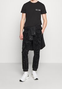 Svart t-shirt med bröstficka och mönstrade svarta sweatpants med en texturerad blommönsterdesign. Matchas med vita sportskor.