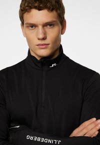 Musta vetoketjullinen huppari, jossa on korkea kaulus. Kaulassa on hillitty logo ja hihassa brändin nimi. Suora, istuva materiaali.