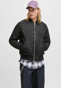 Veste bomber noire avec une finition brillante, poignets et ourlet côtelés, fermeture éclair devant et deux poches latérales. Portée par-dessus une chemise à carreaux avec un jean sombre.