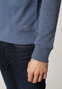 Sudadera azul texturizada con puños y dobladillo acanalados, que presenta una pequeña etiqueta de la marca cerca de la parte inferior. Combinada con jeans de mezclilla oscura.