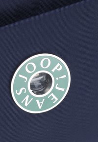 Blå texturerad tyg med en cirkulär metalllogo som har gröna accenter och vit text. Logotypen innehåller en genomskinlig central detalj.