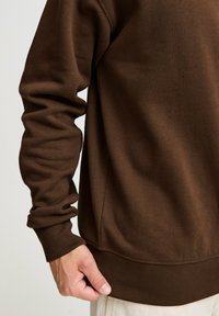 Brun bomullssweater med ribbestrikket mansjetter og hem, med en jevn tekstur og avslappet passform, vist fra siden med en hånd som griper kanten.