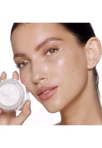 Moisturizing cream in clear hexagonal jar held by a person. The cream is white with a smooth texture. Person has glossy skin and natural makeup.