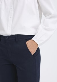 Navyblaue Hose mit Fronttaschen, aus robustem Stoff gefertigt. Ein weißes Hemd mit gerafften Ärmeln ist teilweise sichtbar.