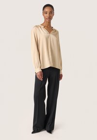 Beige langærmet bluse med V-hals og hætte, der har en subtil tekstur. Sættes sammen med sorte brede bukser og spidse hæle.