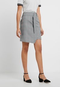 Grijze geruite wikkelrok met een strik in de taille, met een asymmetrische zoom. Gladde stoftextuur, gecombineerd met zwarte puntige hakken.