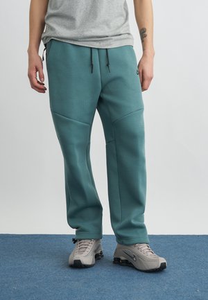 Osoba ubrana w zielone spodnie dresowe, szarą koszulkę i szare buty Nike stoi na niebieskim dywanie na jednolitym tle.