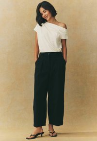 Biela off-shoulder blúzka, voľné čierne nohavice s vysokým pásom a vreckami, a čierne sandále na opätku. Hladký materiál a uvoľnený strih.