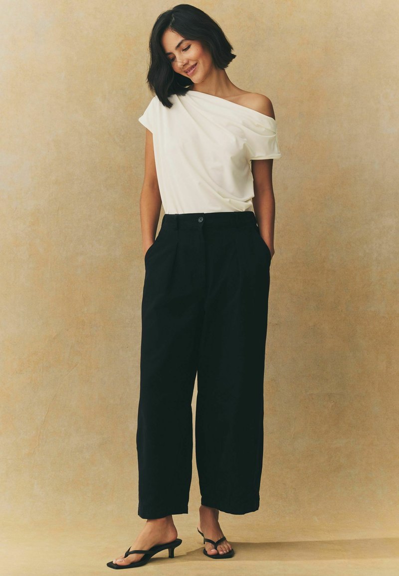 Biela off-shoulder blúzka, voľné čierne nohavice s vysokým pásom a vreckami, a čierne sandále na opätku. Hladký materiál a uvoľnený strih.