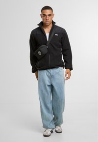 Giacca in pile nera con zip, jeans a gamba larga azzurri, e una piccola borsa a tracolla. Le sneakers presentano dettagli in bianco e nero.