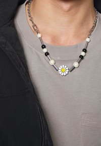 Náhrdelník s černými a bílými korálky, stříbrnými články a bílým přívěskem sedmikrásky se žlutým středem, nošený přes světle šedou košili a černou bundu.