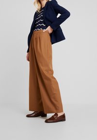 Pantalones de pierna ancha marrones con plisados frontales, acompañados de una blazer azul marino sobre una blusa con patrón de zigzag. Mocasines marrones brillantes completan el conjunto.