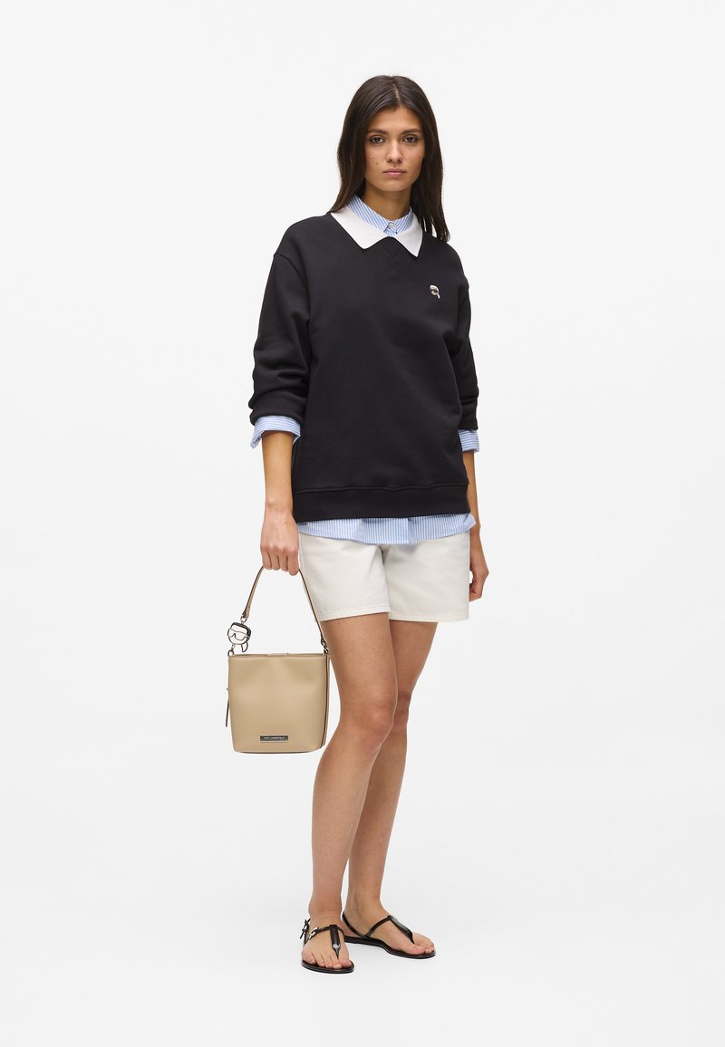 Sweat-shirt noir avec un col blanc sur une chemise à rayures bleues, associé à un short beige et des tongs noires, tenant un sac seau crème.
