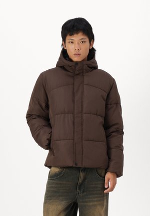 Brun pufferjakke med hætte, der har et quiltet design, trykknaplukning og isolerende polstring. Båret med slidte blå denimbukser.