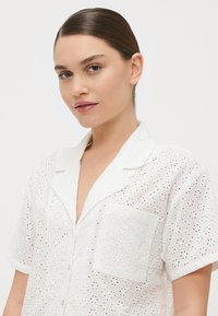 Hvit kortermet skjorte med øyeletstoff, en nothals og brystlomme. Designet har små blomsterutskjæringer over hele skjorten.