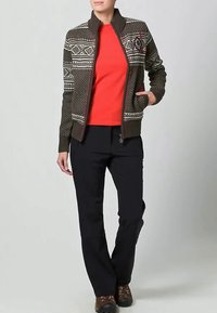 Άτομο που φοράει καρό φούτερ με φερμουάρ πάνω από φωτεινό κόκκινο πουκάμισο, μαύρες παν trousers και καφέ παπούτσια πεζοπορίας, στέκεται με το ένα χέρι στην τσέπη.