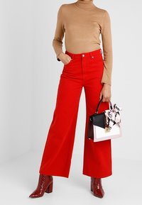 Szerokie czerwone spodnie z bawełny, zestawione z beżowym golfem. W jednej ręce trzyma wielokolorową torebkę ze szalikiem. Całość uzupełniają błyszczące botki za kostkę.