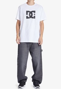 Henkilö, joka wear a white T-shirt with a large black DC logo, loose gray jeans, and black sneakers standing against a white background.