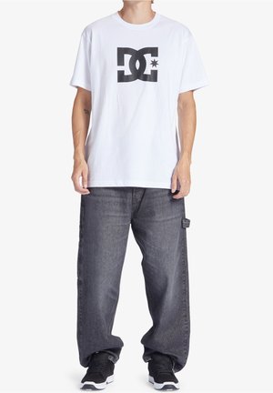 Persona che indossa una maglietta bianca con un grande logo DC nero, jeans grigi larghi e sneakers nere, in piedi contro uno sfondo bianco.