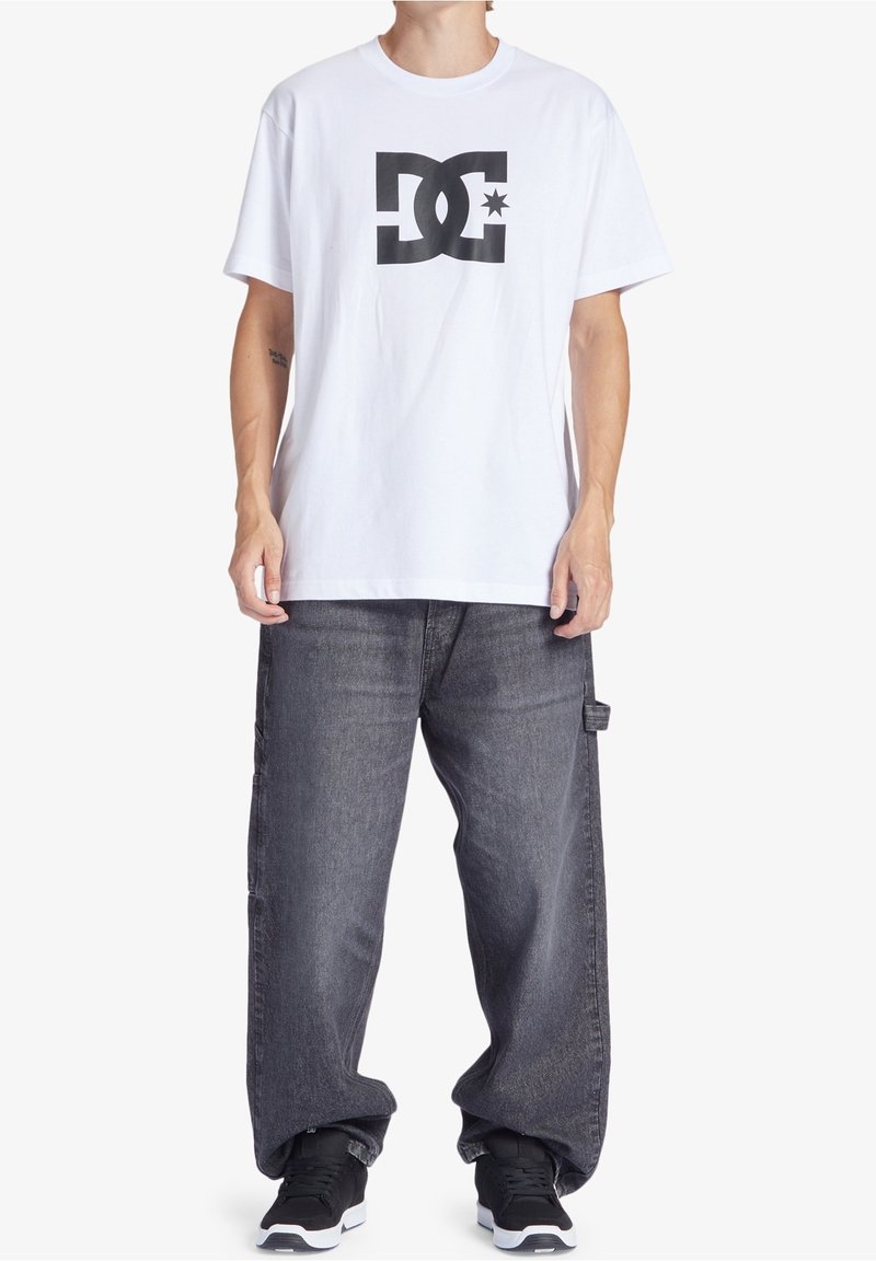 Henkilö, joka wear a white T-shirt with a large black DC logo, loose gray jeans, and black sneakers standing against a white background.
